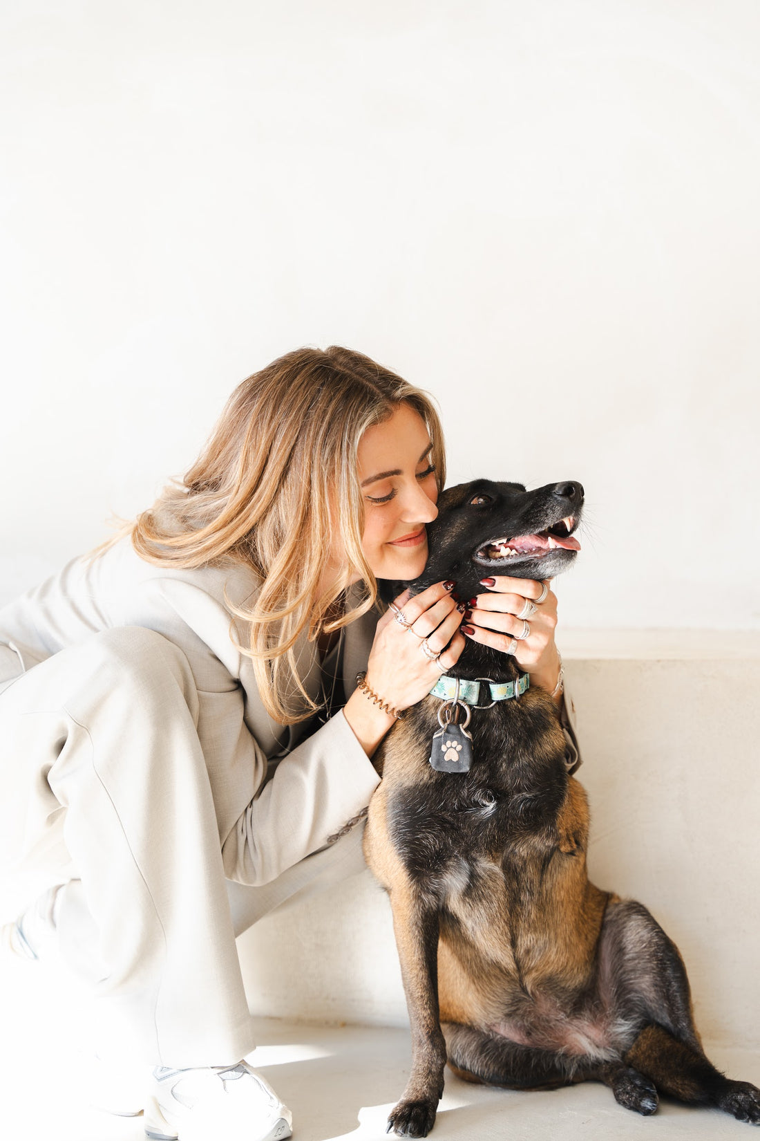 Woman in a beige suit kneels and nuzzles a happy black-and-tan shepherd mix wearing a teal collar.