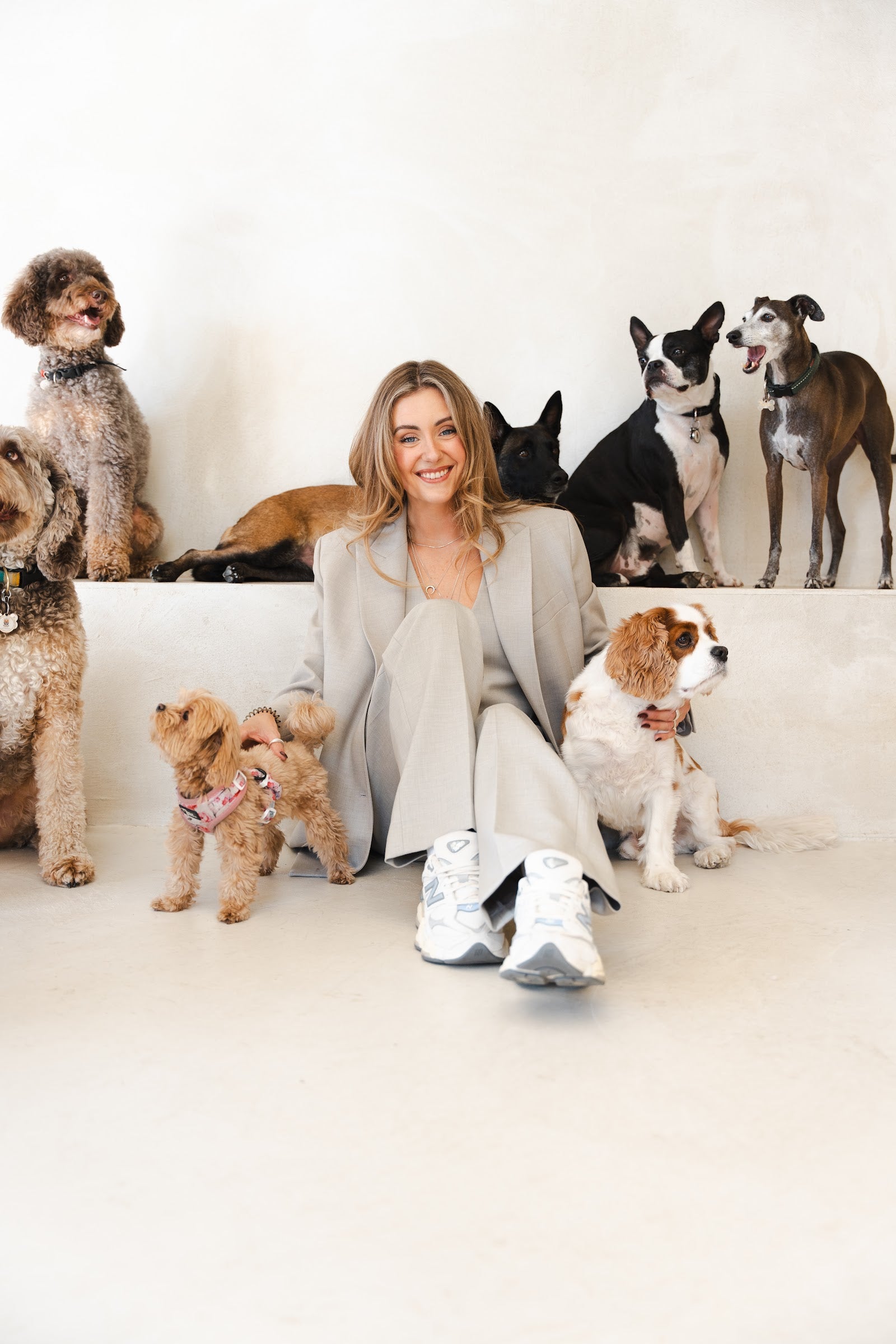 Woman in a beige suit sits on the floor smiling, surrounded by about seven dogs of different breeds on a pale ledge.