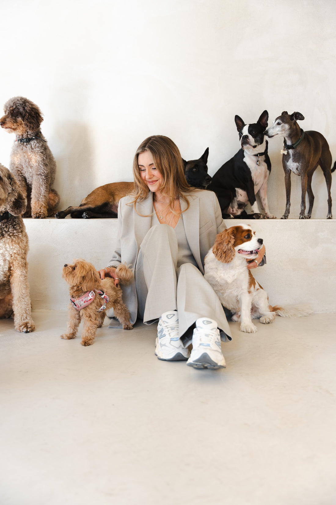 Woman in a beige suit on the floor smiles down at a small apricot dog while several other dogs sit around her.